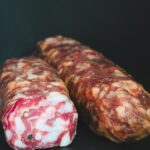 Two rustic cured salami sticks with marbled texture on a dark surface, perfect for meat lovers.
