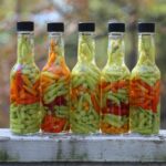 Five bottles filled with vibrant pickled hot peppers displayed outdoors on a sunny day.