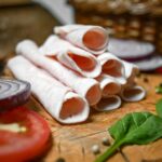 Rolled deli slices on a wooden board with fresh onions and spinach leaves, perfect for culinary presentations.